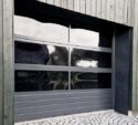 A closed, modern garage door with four glass panels set in a vertical wood-paneled exterior wall; cobblestone driveway in front.