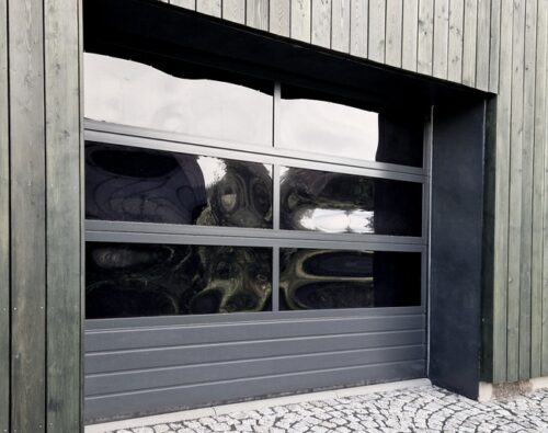 A closed, modern garage door with four glass panels set in a vertical wood-paneled exterior wall; cobblestone driveway in front.