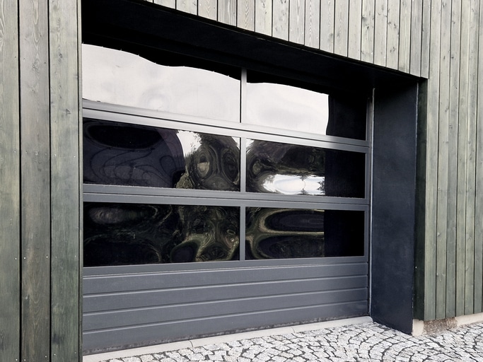 A closed, modern garage door with four glass panels set in a vertical wood-paneled exterior wall; cobblestone driveway in front.