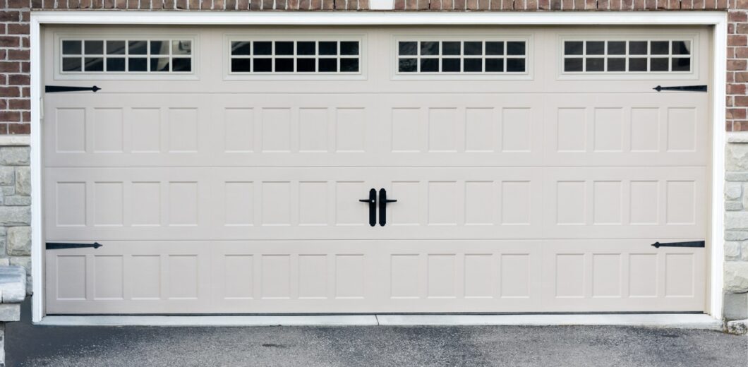 Closed beige garage door with rectangular panels and three small windows at the top, set in a brick and stone exterior wall.