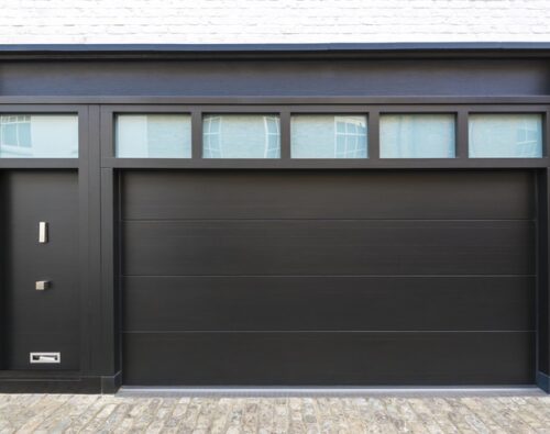 Modern black garage door with four rectangular windows above and an adjacent black entrance door, set in a white brick building with a stone driveway.