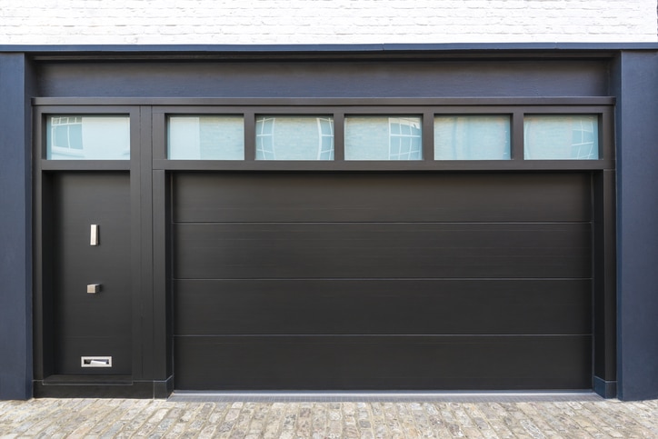 Modern black garage door with four rectangular windows above and an adjacent black entrance door, set in a white brick building with a stone driveway.