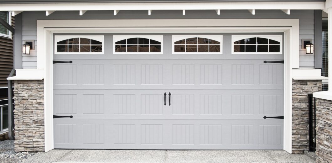 A closed, double-car garage door with vertical panel design, decorative windows, and black hardware on a modern house with stone accents.