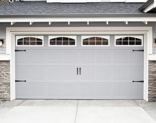A closed, double-car garage door with vertical panel design, decorative windows, and black hardware on a modern house with stone accents.