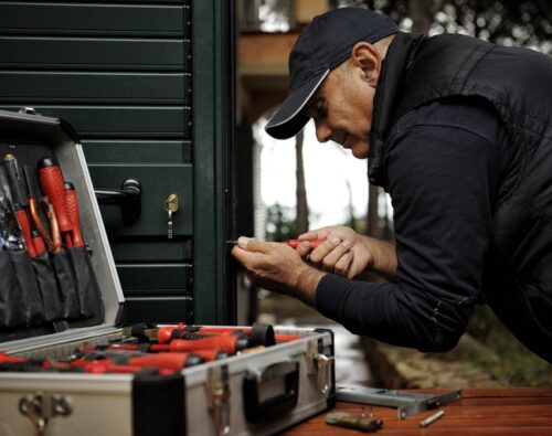 A person in a cap and vest uses a screwdriver to work on a lock, with an open toolbox of various tools nearby.