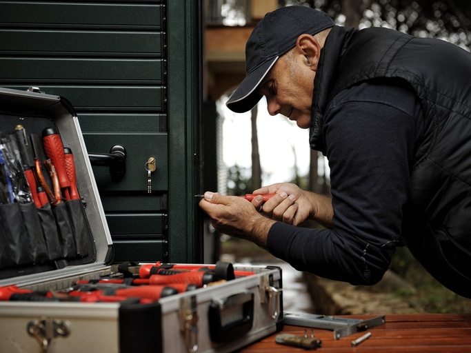 A person in a cap and vest uses a screwdriver to work on a lock, with an open toolbox of various tools nearby.