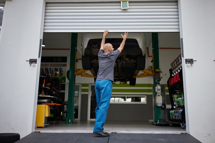 A person in blue pants opens a garage door in an auto repair shop with a car lifted on a hydraulic platform inside.