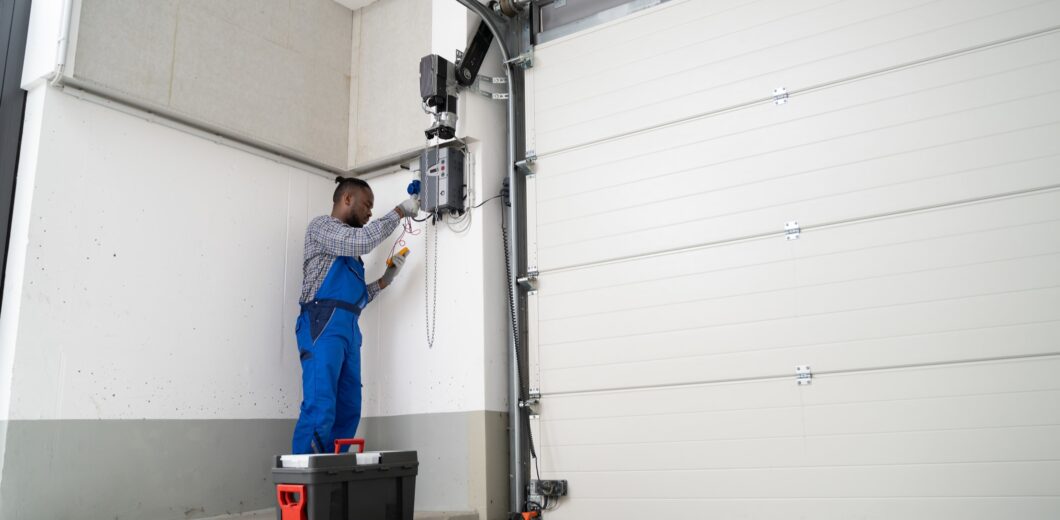 A person wearing blue coveralls repairs or installs a garage door opener using tools, with a toolbox placed on the floor nearby.