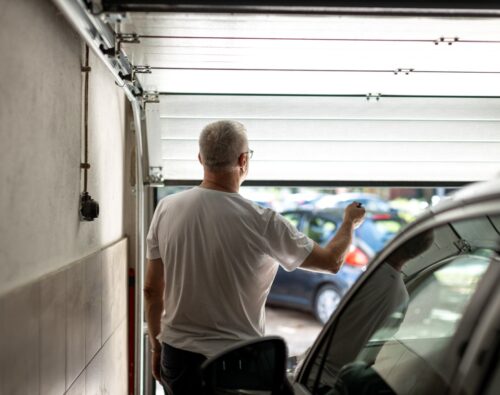 A man with short gray hair stands inside a garage, holding a remote control while opening the garage door. A car is parked inside, and other vehicles are visible outside.