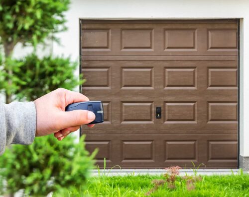 A person’s hand holds a remote control in front of a closed brown garage door, with greenery visible in the foreground.