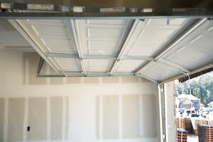 Partially open white garage door inside an unfinished garage with exposed drywall. Exterior view shows houses and construction materials.