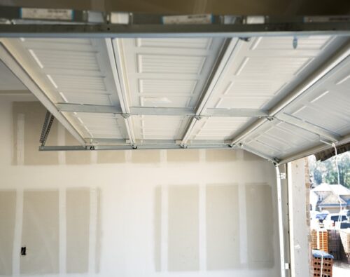 Partially open white garage door inside an unfinished garage with exposed drywall. Exterior view shows houses and construction materials.