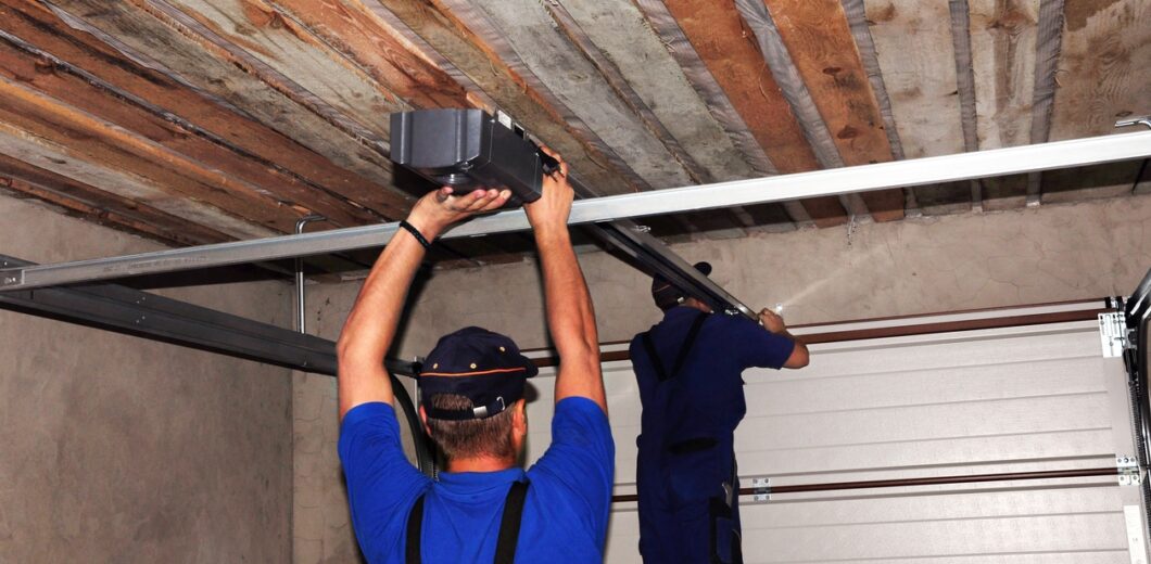 Two workers in blue uniforms install or repair a garage door opener mechanism on the ceiling of a garage with exposed wooden beams.