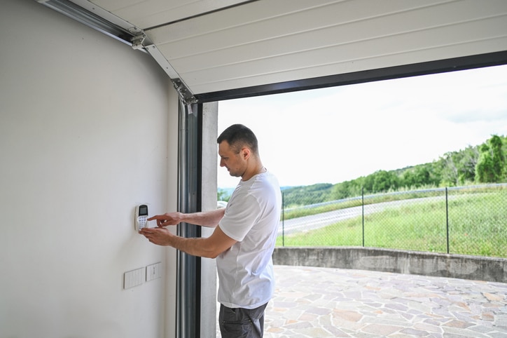 A man in a white shirt stands in a garage, using a keypad mounted on the wall near the open garage door, with a driveway and greenery outside.