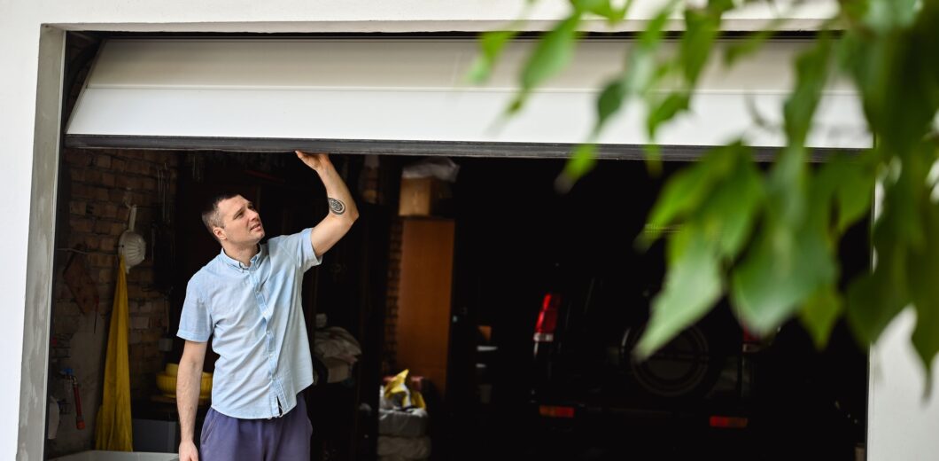 A man in casual clothes lifts a partially open garage door, with a car and various items visible inside the garage; green leaves are in the foreground.