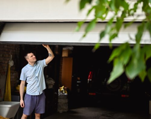 A man in casual clothes lifts a partially open garage door, with a car and various items visible inside the garage; green leaves are in the foreground.