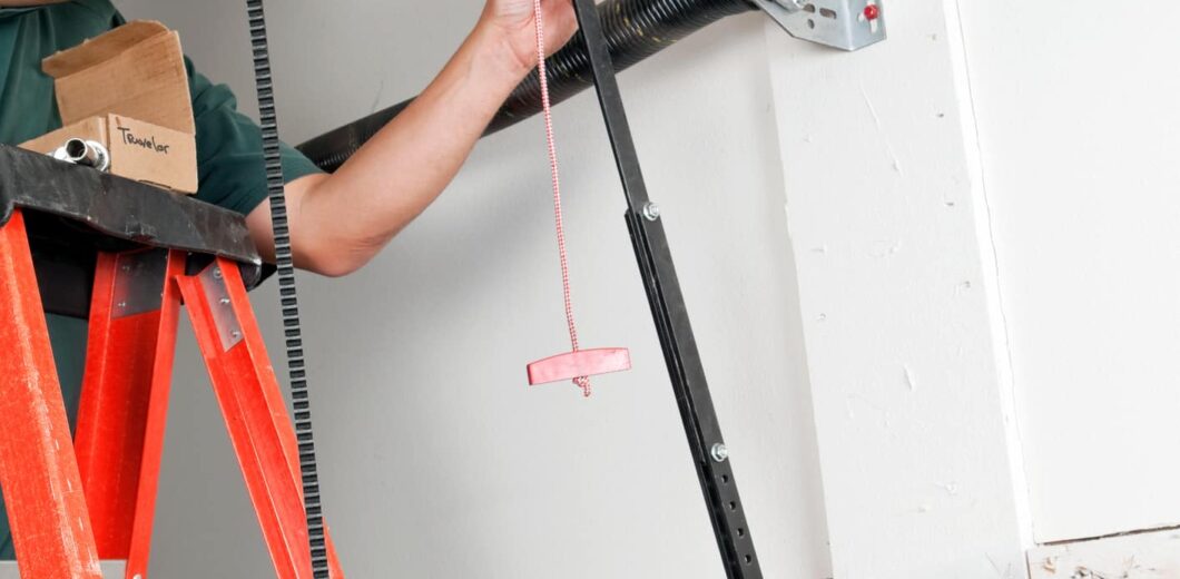 Person on a red ladder repairs or installs a garage door spring mechanism, using tools on the garage ceiling.