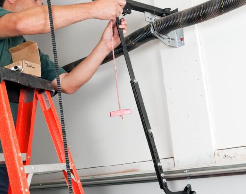Person on a red ladder repairs or installs a garage door spring mechanism, using tools on the garage ceiling.