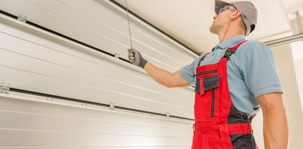 A technician in red overalls inspects or repairs a raised garage door, holding a cable and looking up at the mechanism.
