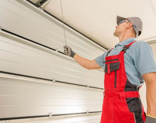 A technician in red overalls inspects or repairs a raised garage door, holding a cable and looking up at the mechanism.