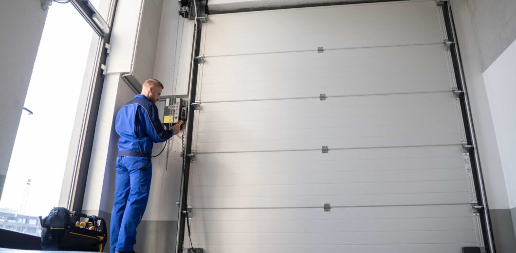 A technician in blue overalls operates a control panel next to a large closed industrial garage door inside a building.