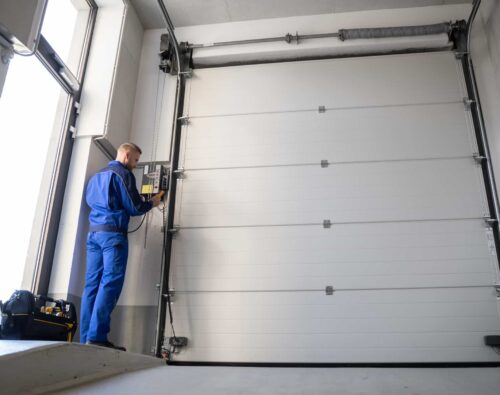 A technician in blue overalls operates a control panel next to a large closed industrial garage door inside a building.