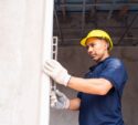 A construction worker in a yellow hard hat and gloves uses a level tool to check a wall's alignment indoors.