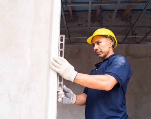 A construction worker in a yellow hard hat and gloves uses a level tool to check a wall's alignment indoors.
