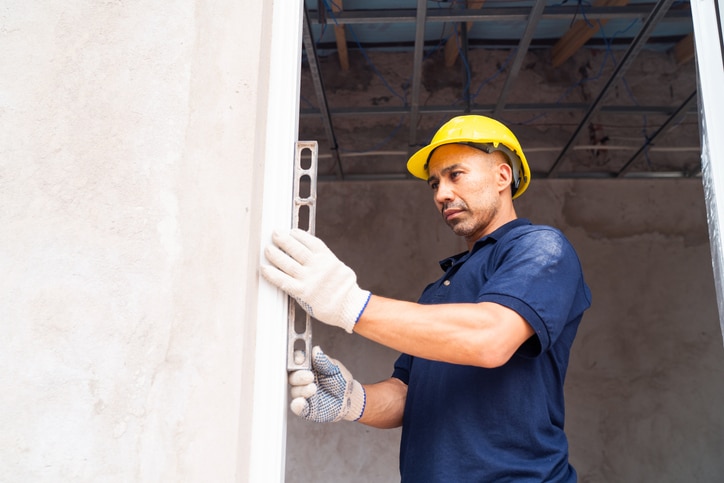 A construction worker in a yellow hard hat and gloves uses a level tool to check a wall's alignment indoors.