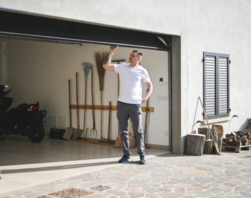 Man in casual clothes standing in front of a half-open garage door, looking up while holding the door; various tools and firewood are visible inside and beside the garage.