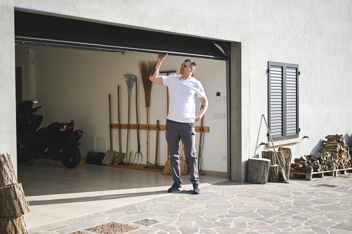 Man in casual clothes standing in front of a half-open garage door, looking up while holding the door; various tools and firewood are visible inside and beside the garage.