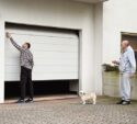 A man opens a garage door while another man stands nearby holding a remote control, and a small dog stands on the driveway.