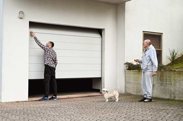 A man opens a garage door while another man stands nearby holding a remote control, and a small dog stands on the driveway.
