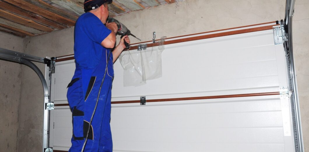 A person in blue overalls stands on a ladder, using a power drill to install or repair a garage door mechanism.