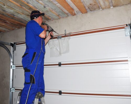 A person in blue overalls stands on a ladder, using a power drill to install or repair a garage door mechanism.