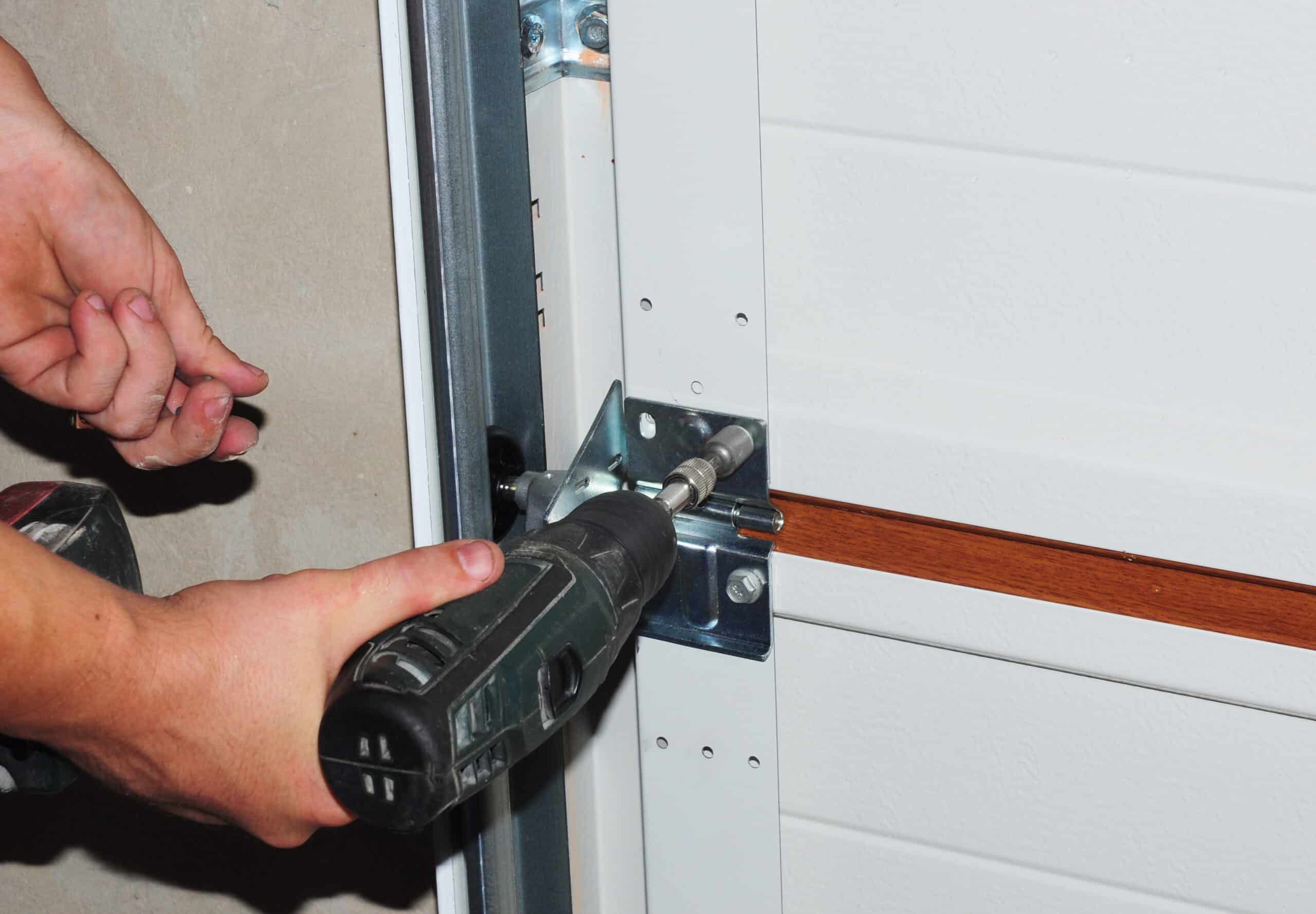 Person using a power drill to fasten or unfasten a screw on a metal bracket attached to a garage door.