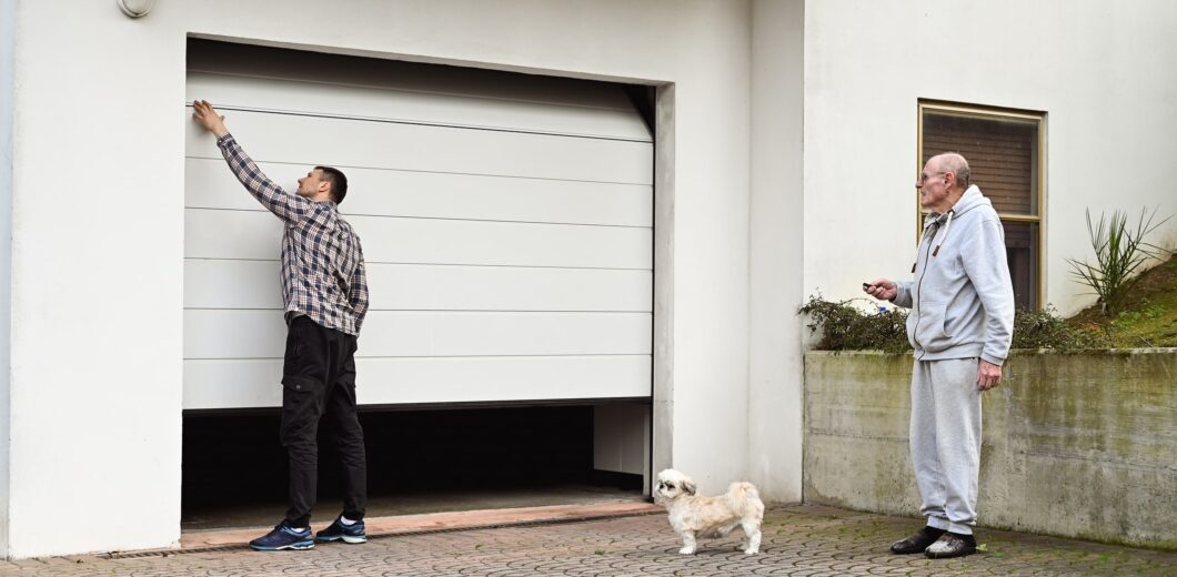 A man in a plaid shirt opens a garage door while another man in gray sweatpants stands nearby holding a remote, with a small dog beside him.