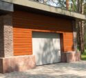A closed garage door on a modern building with wood and stone exterior, surrounded by trees and a paved driveway.