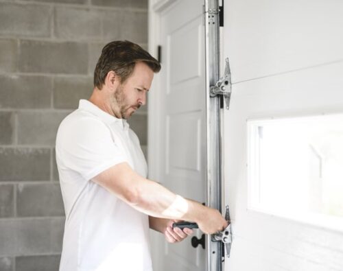 A man in a white shirt uses a tool to adjust the hardware on a white garage door inside a garage with cinderblock walls.