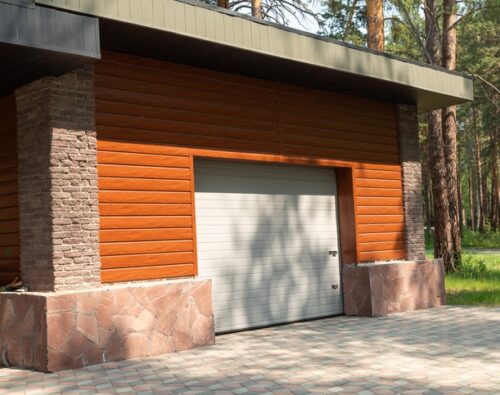 A closed garage door on a modern building with wood and stone exterior, surrounded by trees and a paved driveway.