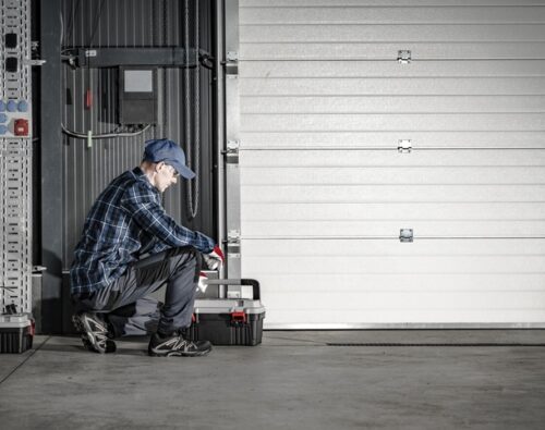 A person in work clothes and a cap kneels beside a closed garage door, working with a toolbox in an industrial setting.