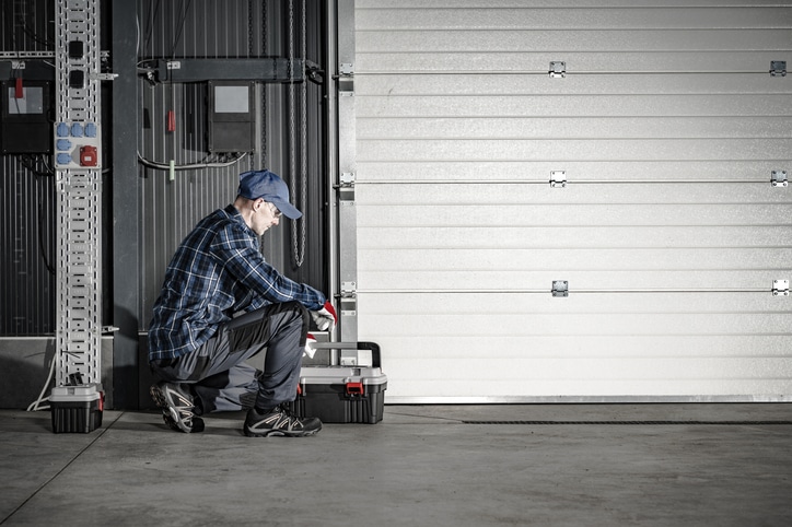 A person in work clothes and a cap kneels beside a closed garage door, working with a toolbox in an industrial setting.