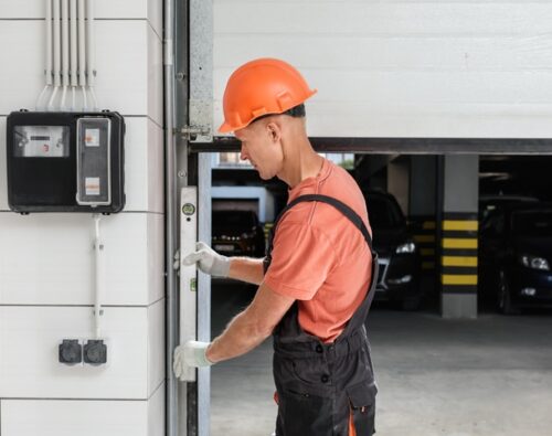 A worker in an orange hard hat and gloves is inspecting or repairing a garage door in a parking garage.