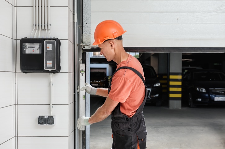 A worker in an orange hard hat and gloves is inspecting or repairing a garage door in a parking garage.