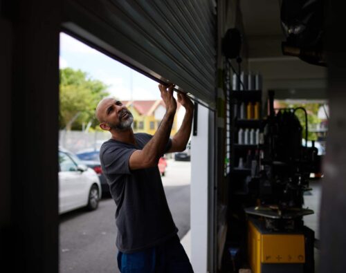 A man in a dark t-shirt pulls open a metal garage door at an automotive repair shop during the day. Cars and buildings are visible outside.