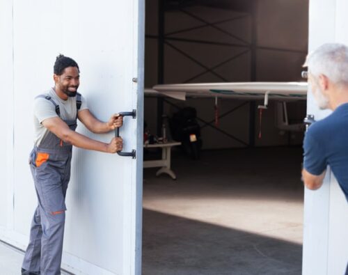 Two men open large hangar doors, revealing a small aircraft inside.
