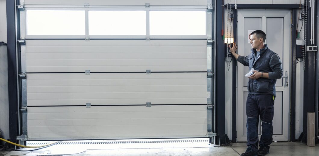 A man in work clothes operates a control on the wall to open or close a large industrial garage door inside a well-lit facility.