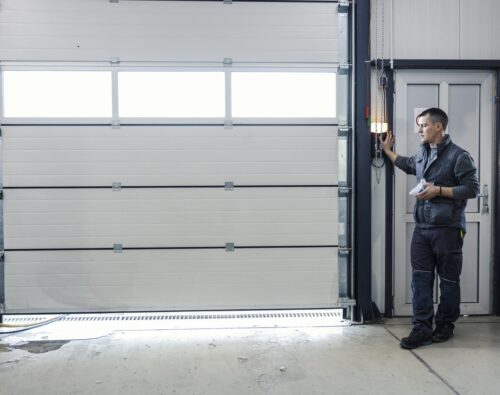 A man in work clothes operates a control on the wall to open or close a large industrial garage door inside a well-lit facility.
