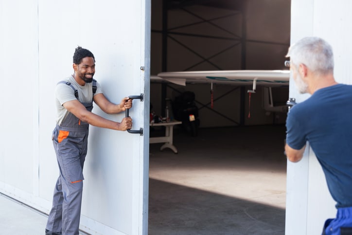 Two men open large hangar doors to reveal the nose of a small airplane inside.
