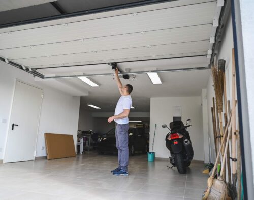 A person stands in a garage opening an overhead door by hand, with cars, a scooter, tools, and cleaning supplies visible inside.